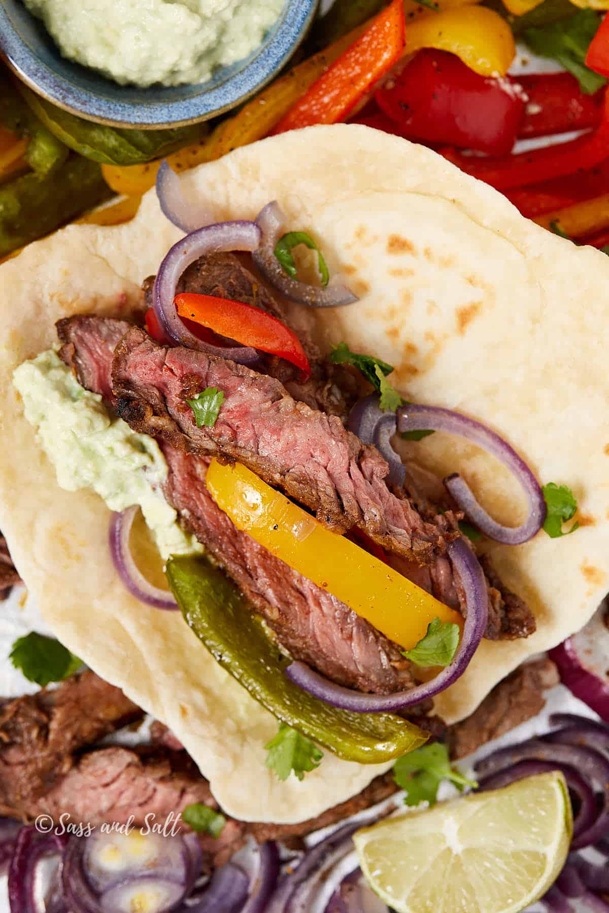 A close-up of a beef fajita on a tortilla, topped with colorful bell peppers, red onion slices, and cilantro. A dollop of avocadeo sauce sits beside, with a lime wedge nearby. Sautéed peppers are visible in the background.