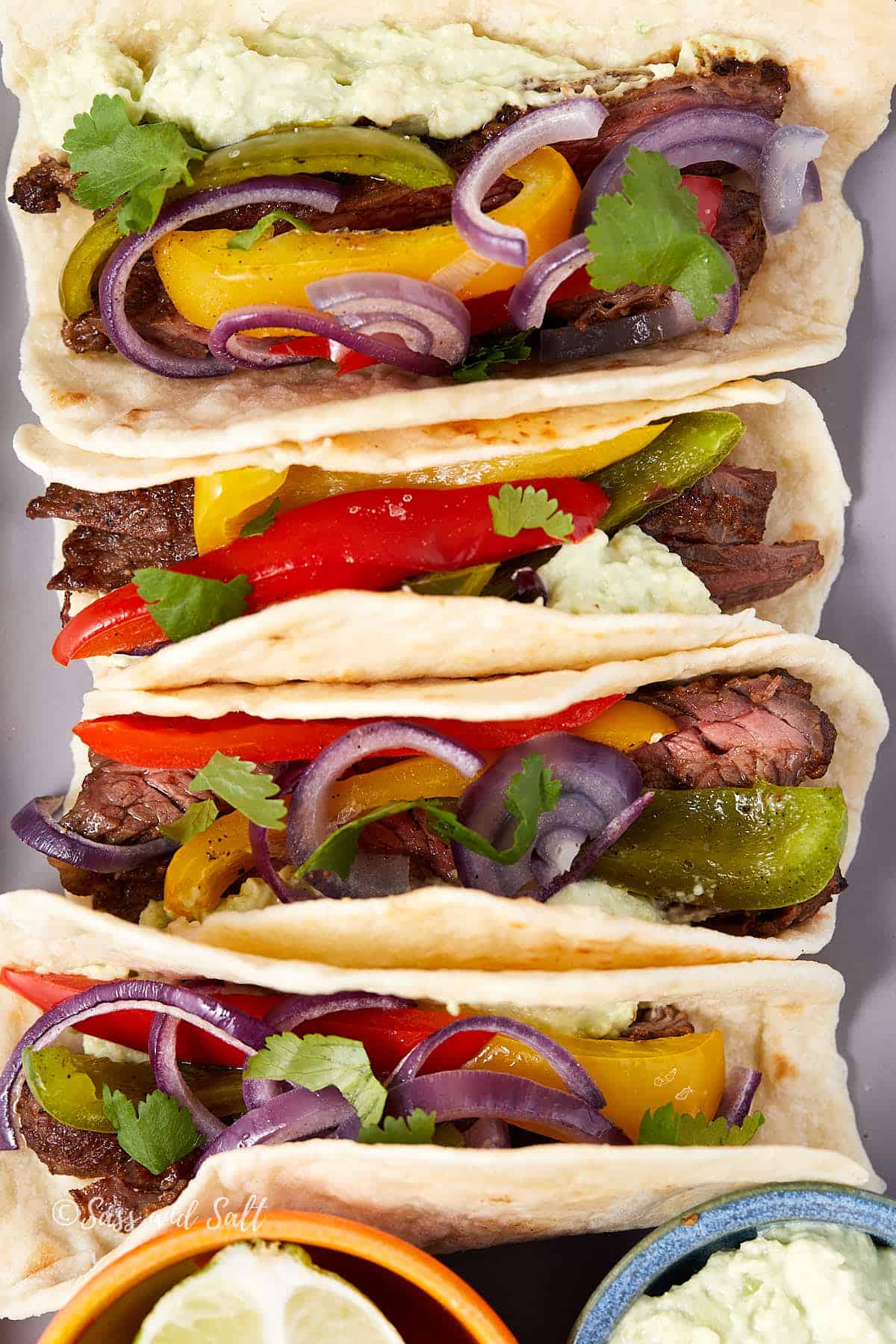 4 skirt steak fajitas on flour tortillas with roasted peppers, red onion, cilantro, and avocado cream, with a lime wedge and a small bowl of avocado cream on the side.
