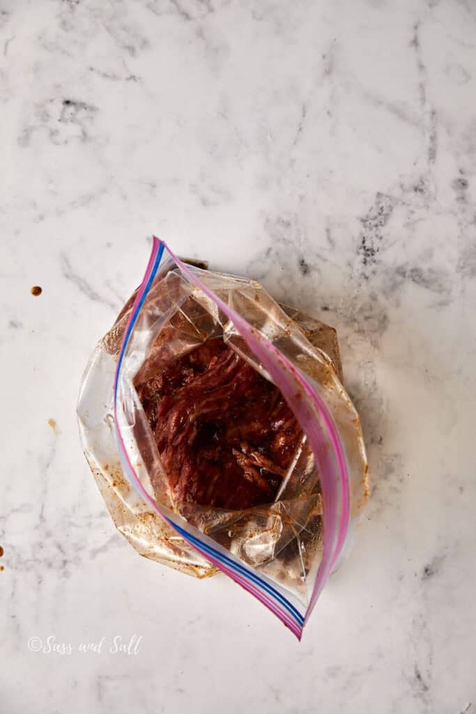 A plastic zipper bag filled with marinating pieces of meat sits on a marble countertop. The marinade appears dark and rich, contrasting with the light surface.