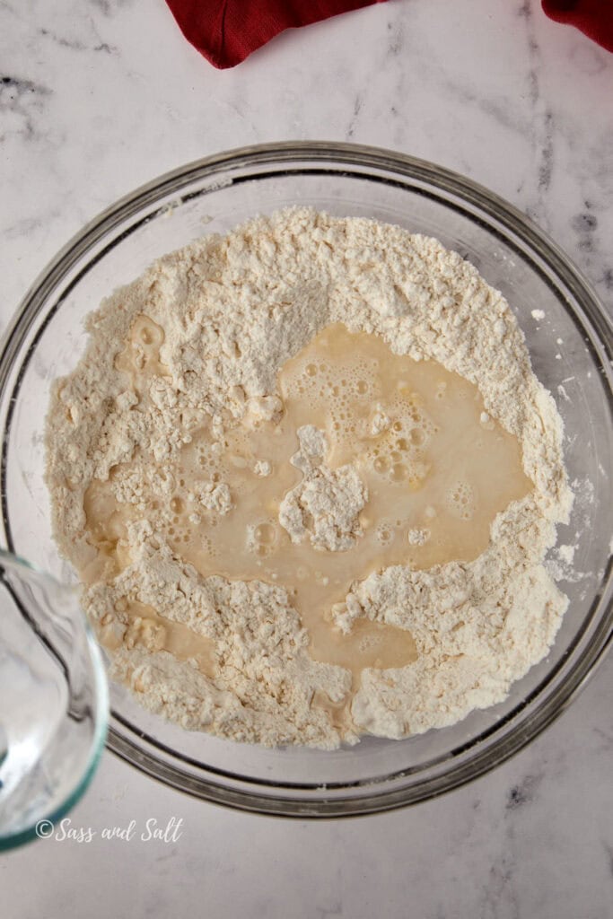 A glass bowl contains a mixture of flour and liquid, ready for mixing. It is set on a marble countertop. A glass pitcher is visible on the left, suggesting it was used to pour the liquid. A red cloth partially shows at the top edge.