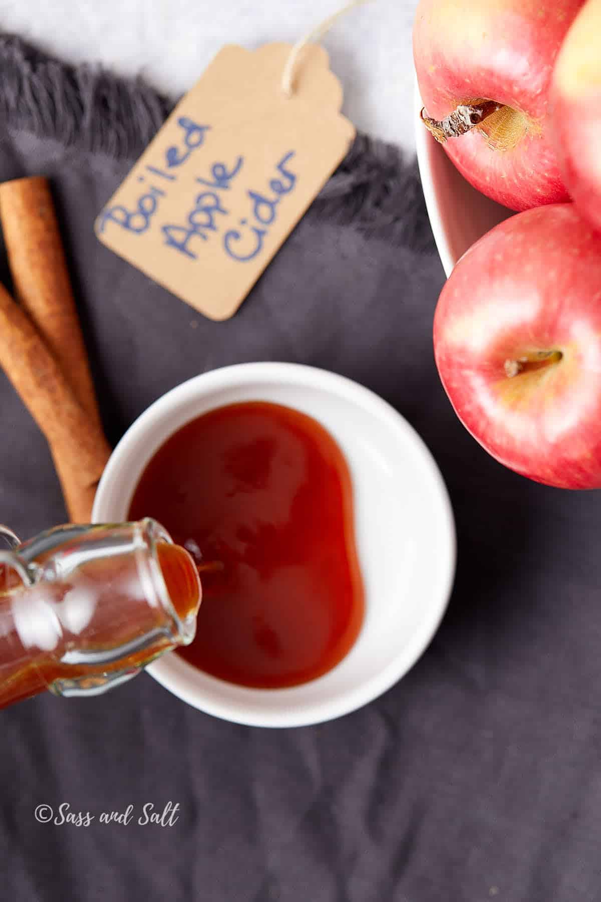 A glass bottle is pouring boiled apple cider into a small white dish. Beside it are red apples in a bowl, cinnamon sticks, and a brown tag labeled "Boiled Apple Cider" on a dark cloth.