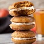 A stack of three pumpkin spice whoopie pies with cream filling. The top one is bitten, revealing the fluffy interior. A blurred glass of orange liquid and peaches are in the background.