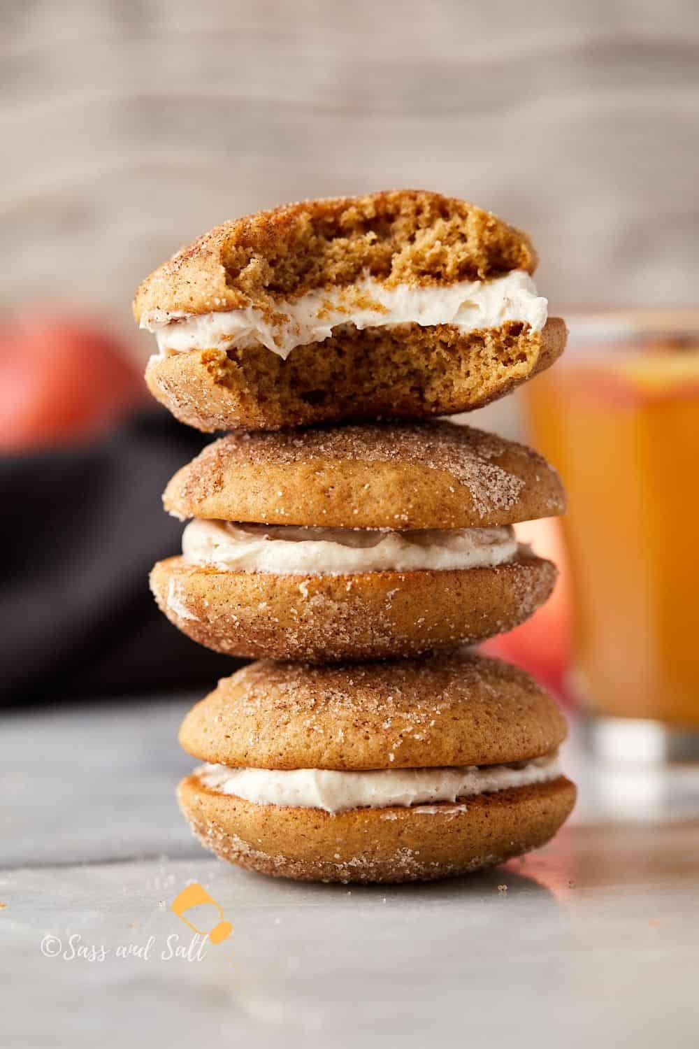 A stack of three pumpkin spice whoopie pies with cream filling. The top one is bitten, revealing the fluffy interior. A blurred glass of orange liquid and peaches are in the background.