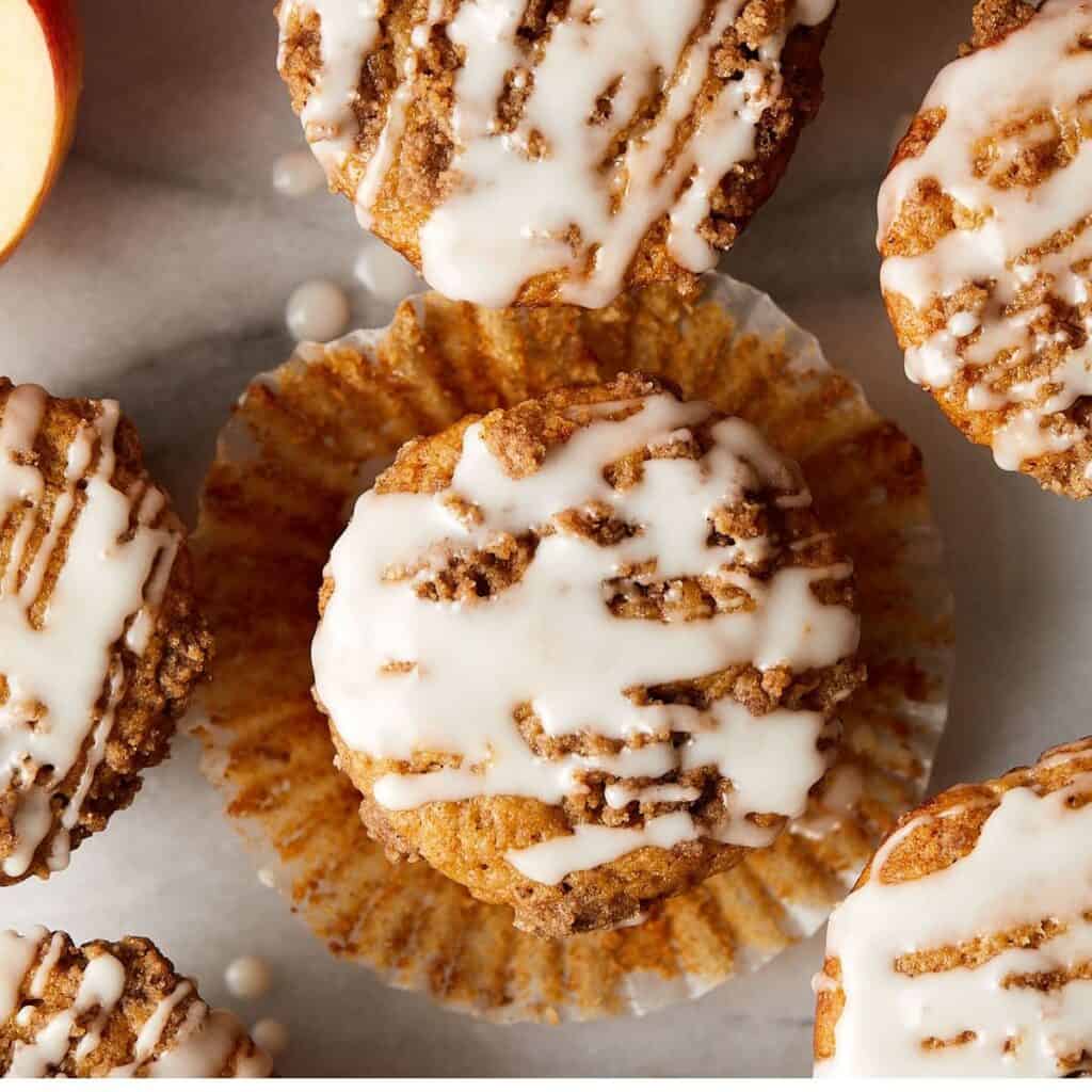 Overhead view of baked muffins with a crumbly topping and white glaze drizzle, one muffin partly unwrapped from its paper liner on a light-colored surface.