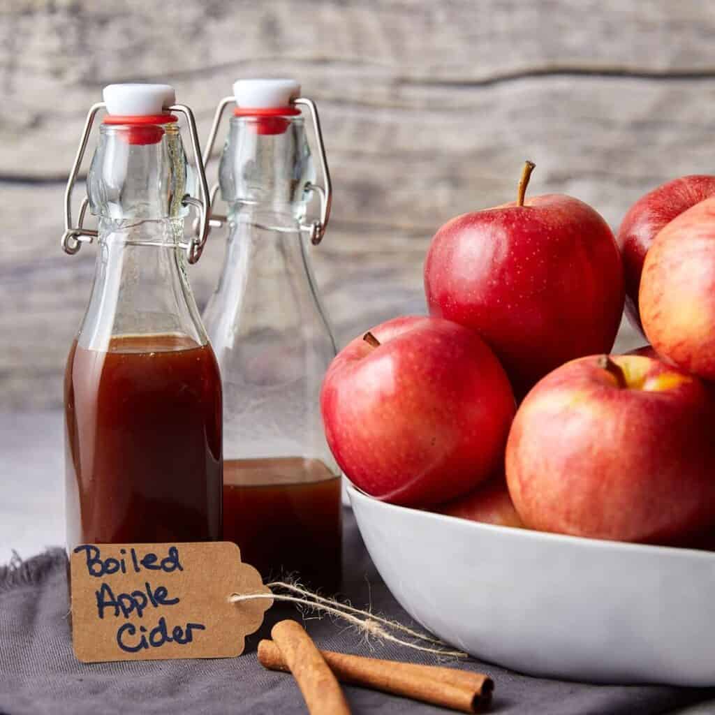 Two glass bottles, one filled with brown liquid labeled "Boiled Apple Cider," stand on a cloth with a cinnamon stick. A white bowl filled with red apples is beside them, set against a rustic wooden background.