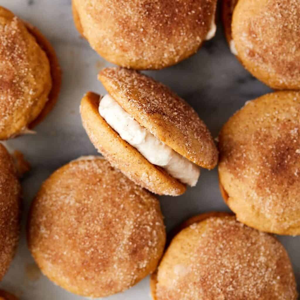 A close-up of pumpkin whoopie pies with cream filling. The cookies are dusted with cinnamon sugar, giving a textured appearance. One pie is slightly open, revealing the creamy filling inside. The background is a light-colored surface.