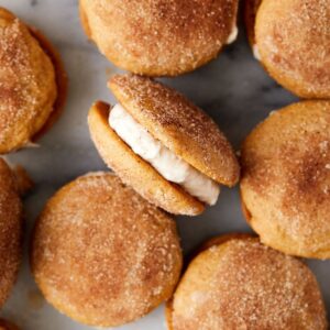 A close-up of pumpkin whoopie pies with cream filling. The cookies are dusted with cinnamon sugar, giving a textured appearance. One pie is slightly open, revealing the creamy filling inside. The background is a light-colored surface.