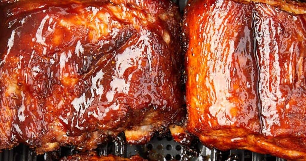 Close-up of juicy, grilled ribs coated in a glossy, dark barbecue sauce. The meat is charred slightly around the edges, highlighting its tenderness and rich flavor, displayed on a black grill surface.