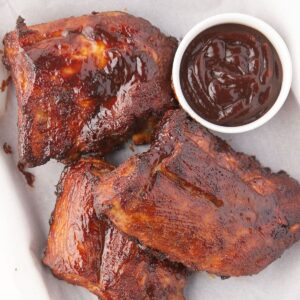Three pieces of glazed barbecue ribs with a small cup of dark barbecue sauce on a white plate. The ribs appear juicy and charred, with a caramelized outer layer.