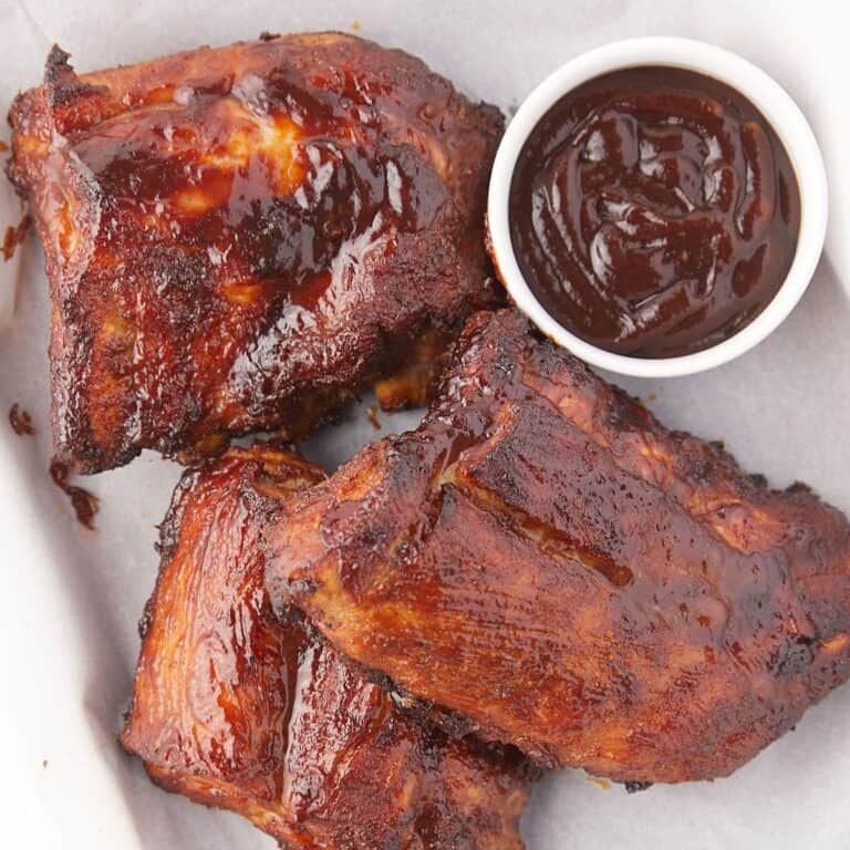 Three pieces of glazed barbecue ribs with a small cup of dark barbecue sauce on a white plate. The ribs appear juicy and charred, with a caramelized outer layer.