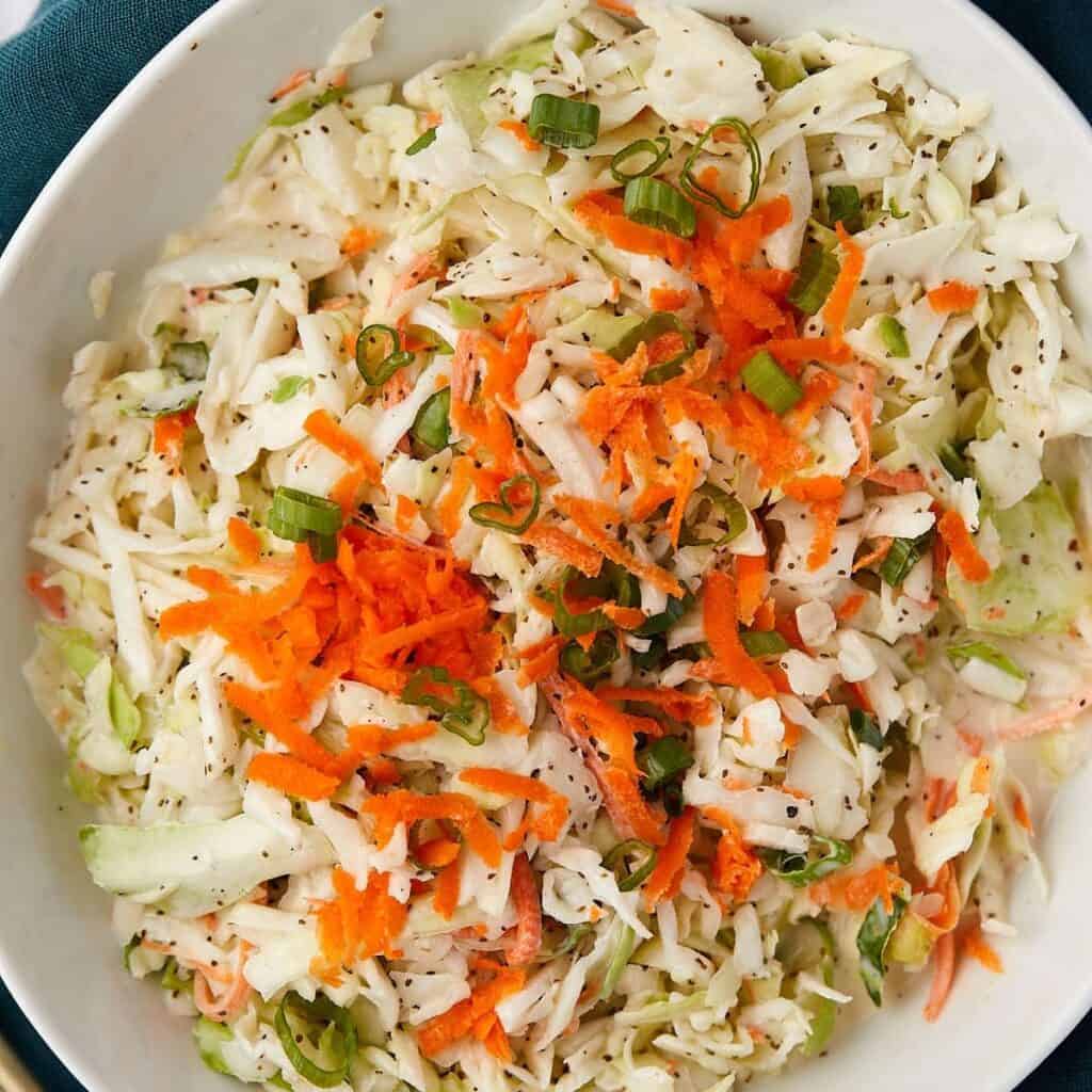 A bowl of coleslaw featuring shredded cabbage, carrots, and sliced green onions. The ingredients are mixed and lightly seasoned, creating a colorful and fresh appearance.