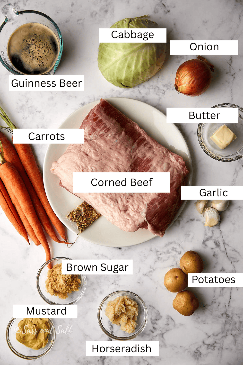 A marble countertop displays ingredients for a corned beef dish: a slab of corned beef, a head of cabbage, carrots, an onion, garlic, potatoes, butter, brown sugar, mustard, horseradish, Guinness beer, and seasonings.