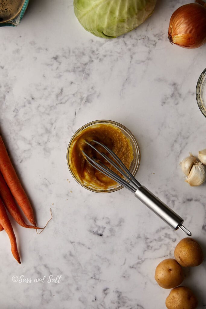 A bowl of brown sauce with a whisk on a marble surface. Surrounding vegetables include carrots, onions, garlic, potatoes, and a cabbage.