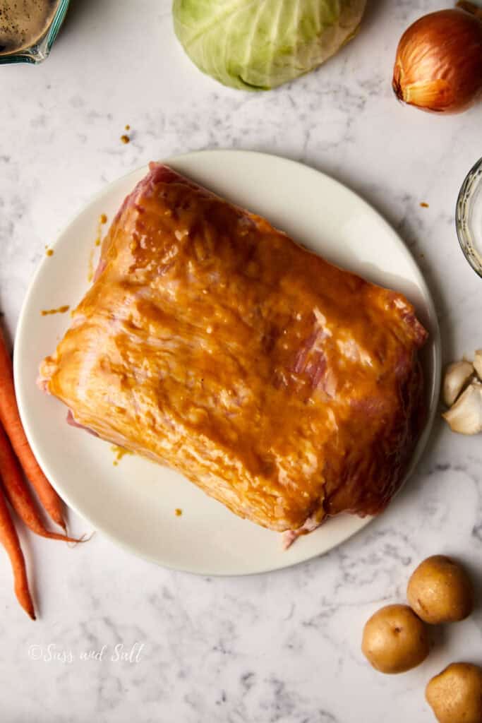 A piece of pork on a white plate, coated with a mustard marinade. Surrounding it are carrots, potatoes, cabbage, an onion, and garlic on a marble countertop.