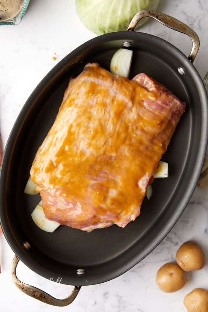 A piece of corned beef coated with mustard sits on a bed of chopped onions in an oval roasting pan. Surrounding the pan are whole potatoes and a cabbage on a marble surface.