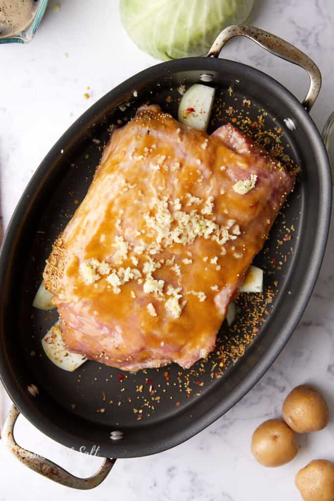 A piece of corned beef covered with mustard and brown sugar glaze in a roasting pan, resting on chunks of onions. Surrounding the pan are ingredients like cabbage and small potatoes on a marble countertop.