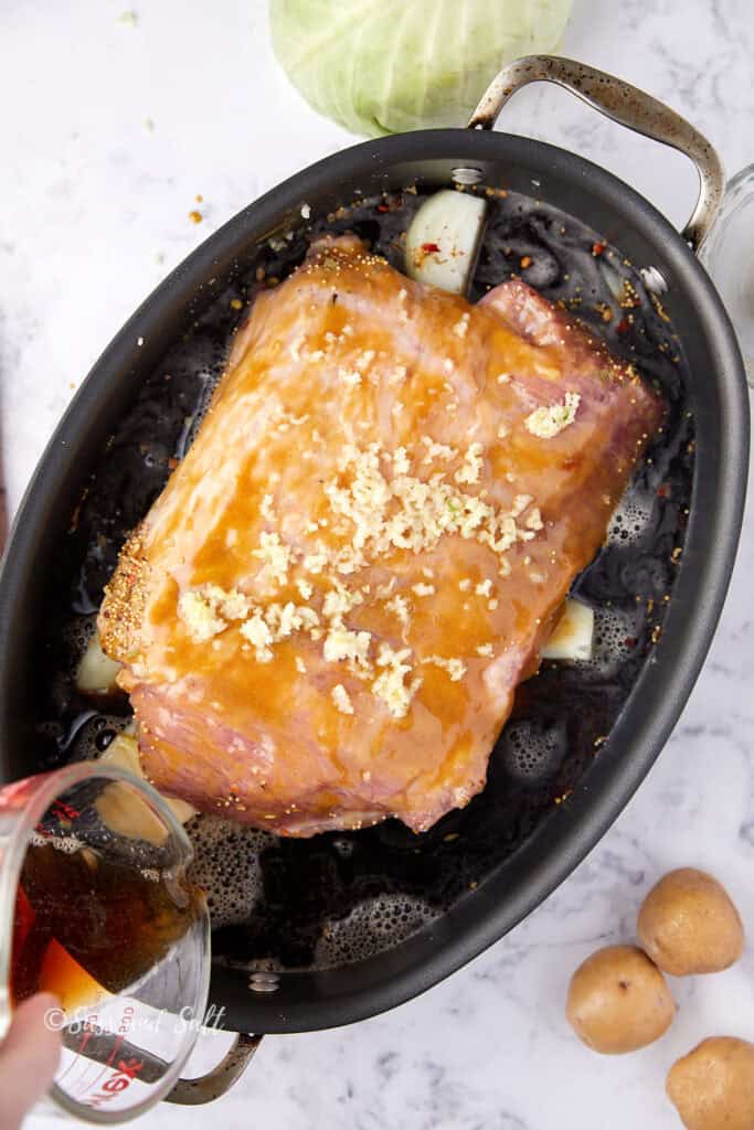 A glazed corned beef brisket in a roasting pan is topped with minced garlic. It's surrounded by onions in a dark broth. A hand is pouring liquid from a measuring cup into the pan. There are small potatoes and a cabbage nearby.