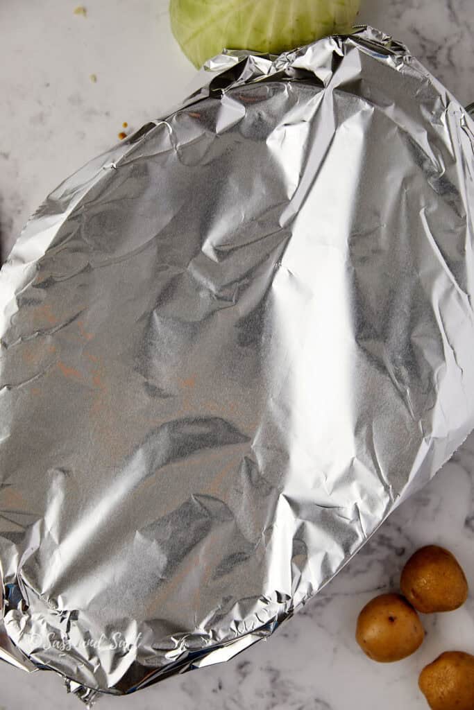 A large dish covered with foil rests on a marble countertop, partially surrounded by a green cabbage and small brown potatoes.
