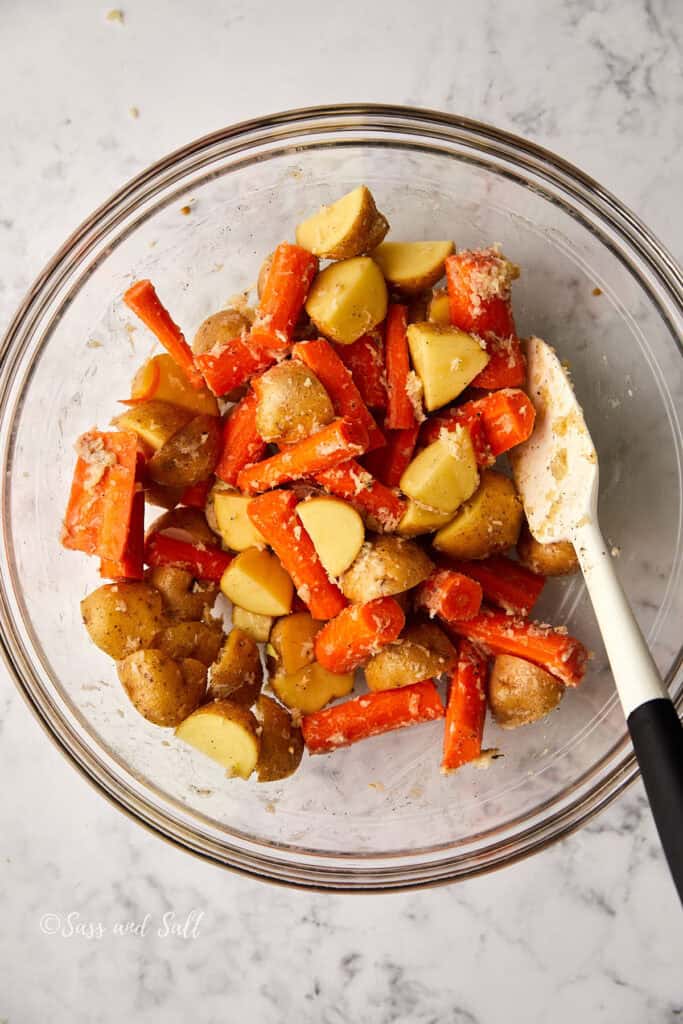 A glass bowl containing chopped potatoes and carrot sticks mixed with seasoning. A white spatula with a black handle is resting inside the bowl on a marble surface.