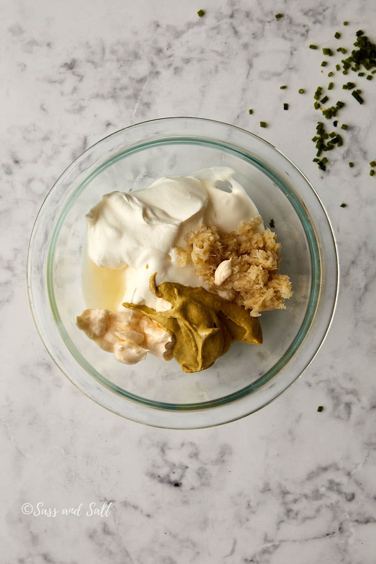 A glass bowl on a marble surface contains a dollop of sour cream, mustard, grated horseradish, and a few whole peeled shrimp. A small sprinkle of chopped herbs is near the bowl.
