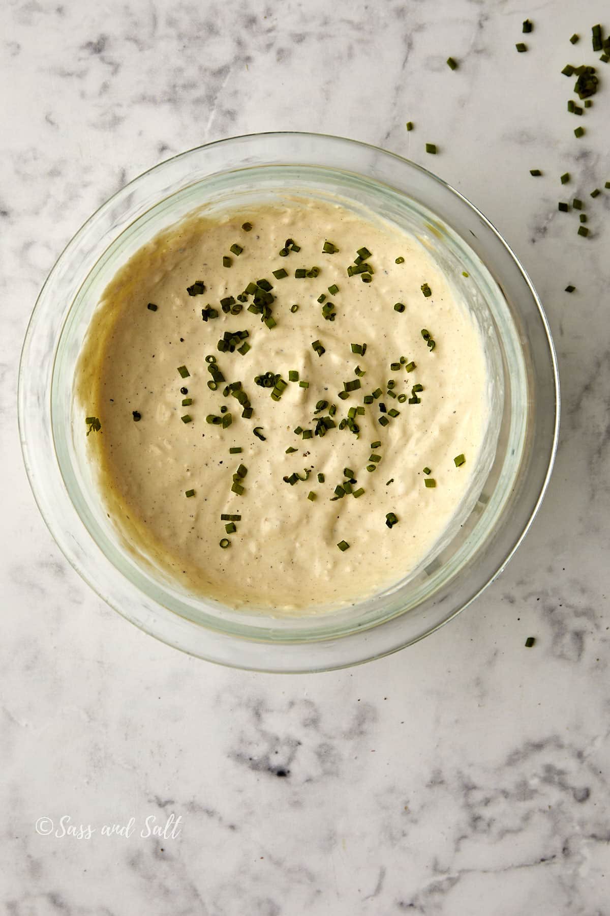 A glass bowl filled with creamy horseradish cream sauce, topped with chopped chives, sits on a marble surface. Small bits of chives are scattered around the bowl.