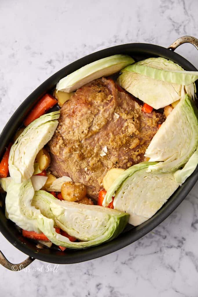 A roasting pan contains a seasoned roast surrounded by wedges of cabbage, potatoes, carrots, and onions. The dish sits on a marble countertop, and the vegetables are arranged neatly around the meat.