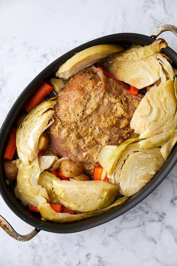 A baked dish in an oval pan with sliced cabbage, potatoes, and carrots surrounding a seasoned meat roast. The vegetables are arranged around the meat, and the composition is set on a marble surface.