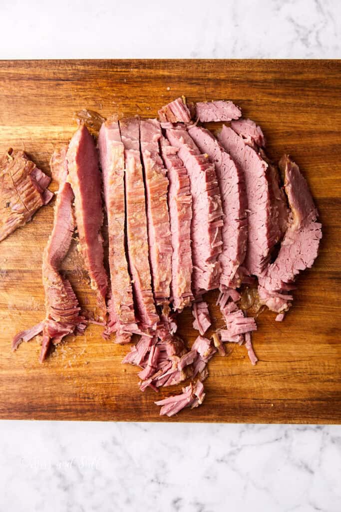 Sliced corned beef brisket arranged on a wooden cutting board, with visible pinkish interior and brown crust. The meat is thinly sliced and neatly organized on a marble surface.