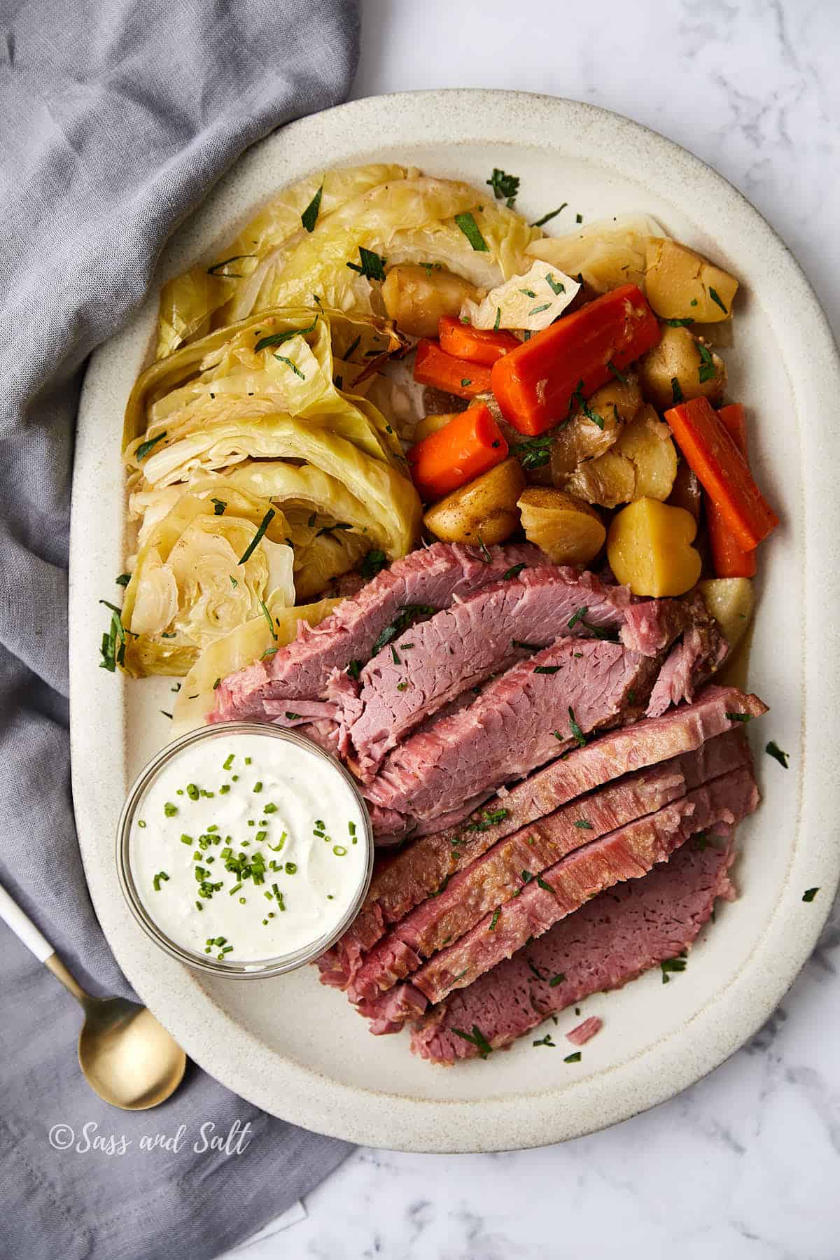 A platter of sliced corned beef, cooked cabbage, carrots, and potatoes, garnished with herbs. A small bowl of creamy white sauce is on the side. The dish is served on a white plate with a gray napkin and a gold spoon nearby.