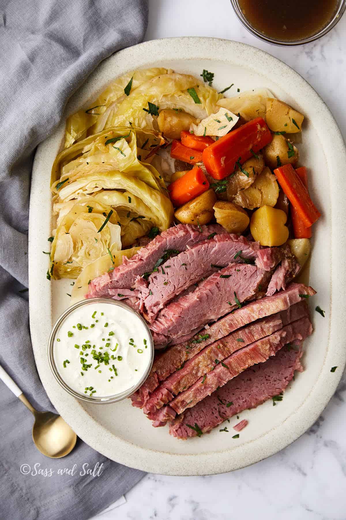 A plate of sliced corned beef, boiled cabbage wedges, carrots, and potatoes. A small bowl of creamy chive sauce is on the side. The dish is on a white plate, with a gray napkin and spoon nearby, set on a marble surface.