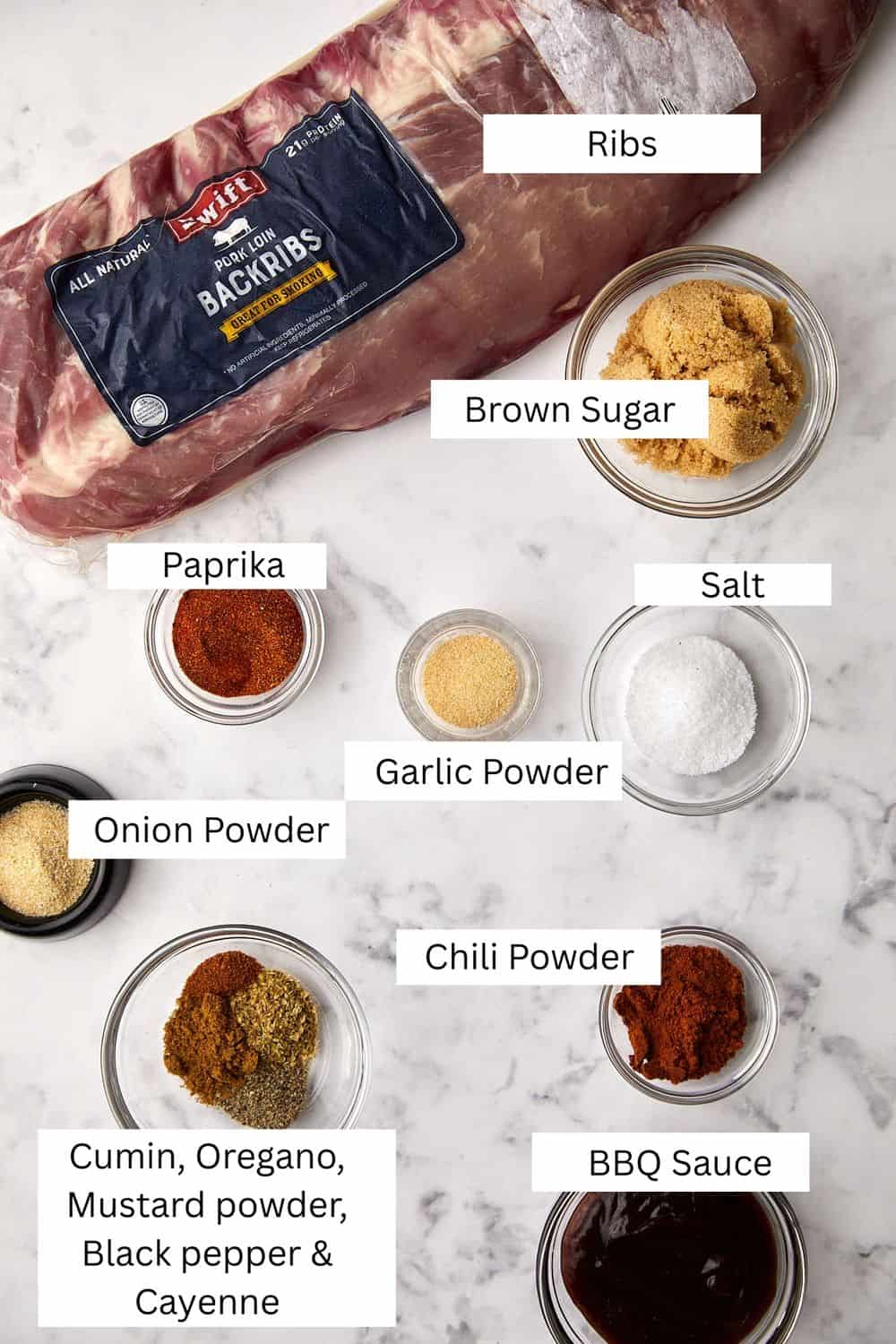 Ingredients laid out on a marble surface for making ribs. Includes ribs in packaging, brown sugar, salt, paprika, onion powder, garlic powder, chili powder, cumin, oregano, mustard powder, black pepper, cayenne, and BBQ sauce.