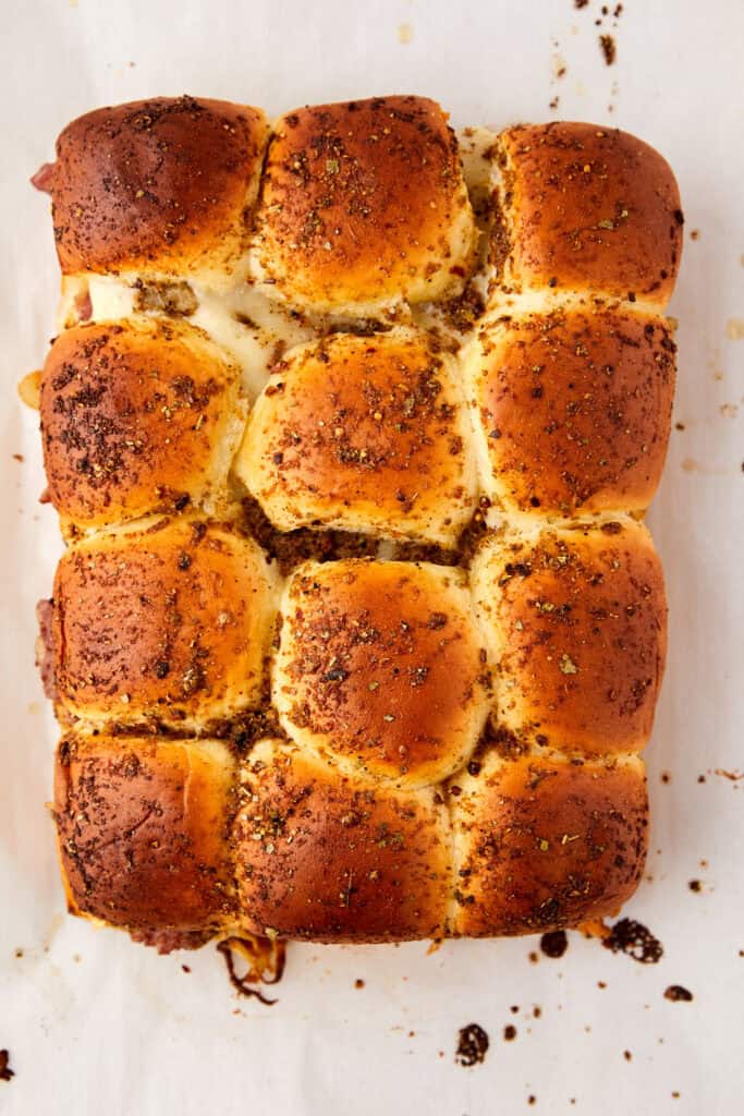 Golden-brown Hawaiian rolls topped with herbs and spices, arranged in a rectangular shape on parchment paper with slightly melted cheese visible between the rolls.