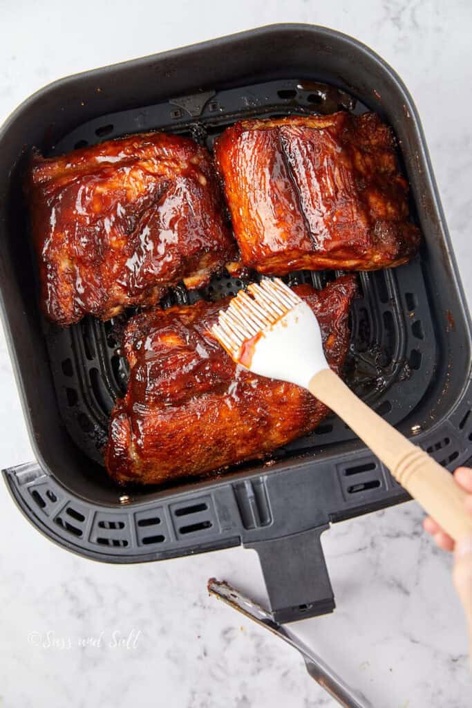 Three pieces of glazed ribs inside an air fryer basket on a marble countertop. A hand is brushing sauce onto the ribs with a white brush.