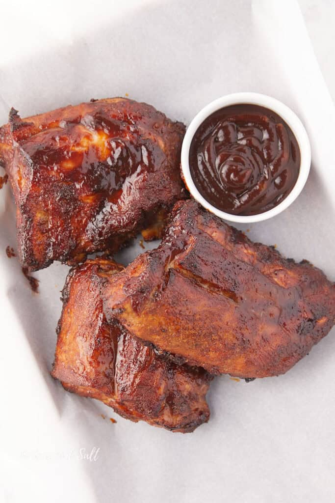 Three pieces of glazed barbecue ribs on a white surface with a small bowl of barbecue sauce beside them.