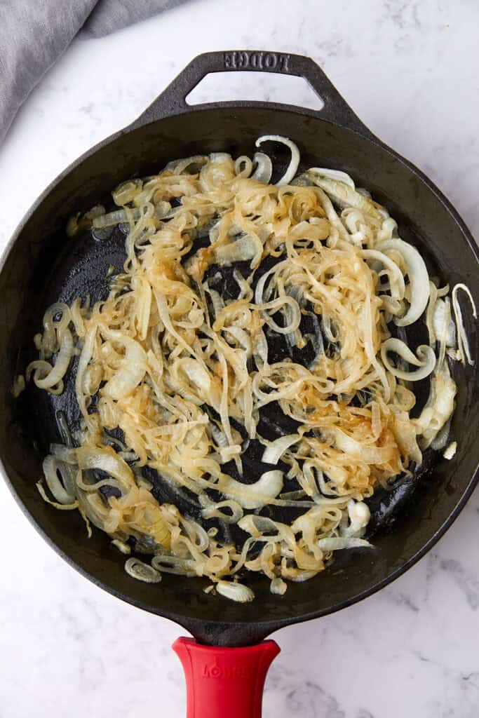 Caramelized onions cooking in a black cast iron skillet with a red handle, sitting on a white marble surface. Some onions are golden brown while others are still light in color.