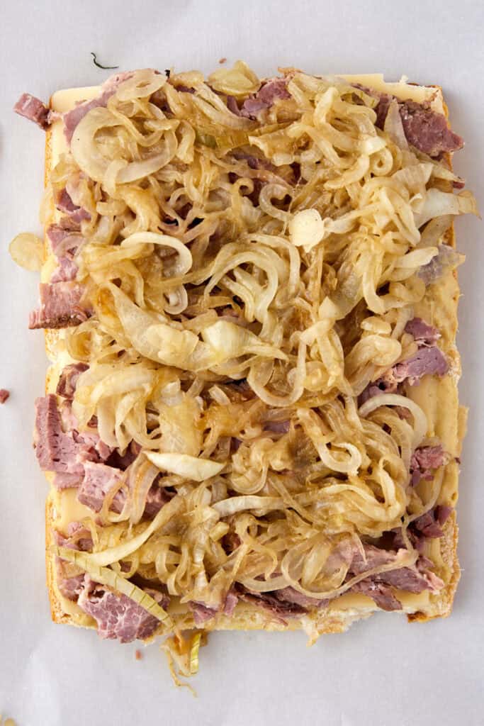 An overhead view of an open-faced sandwich topped with sliced cooked onions and pieces of beef on a rectangular slice of bread, set on white parchment paper.