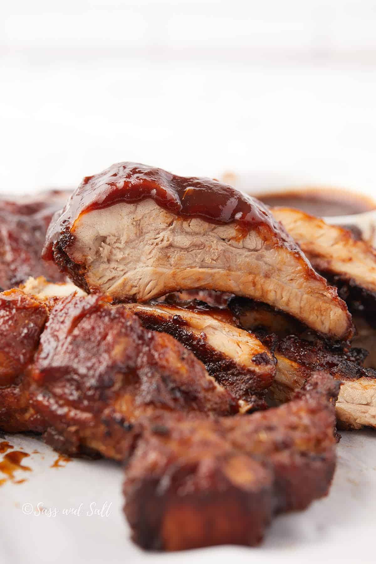Close-up of juicy, sliced barbecued ribs glazed with a shiny layer of barbecue sauce. The meat appears tender and well-cooked, with a visible layer of seasoning and sauce. Additional slices are piled in the background.