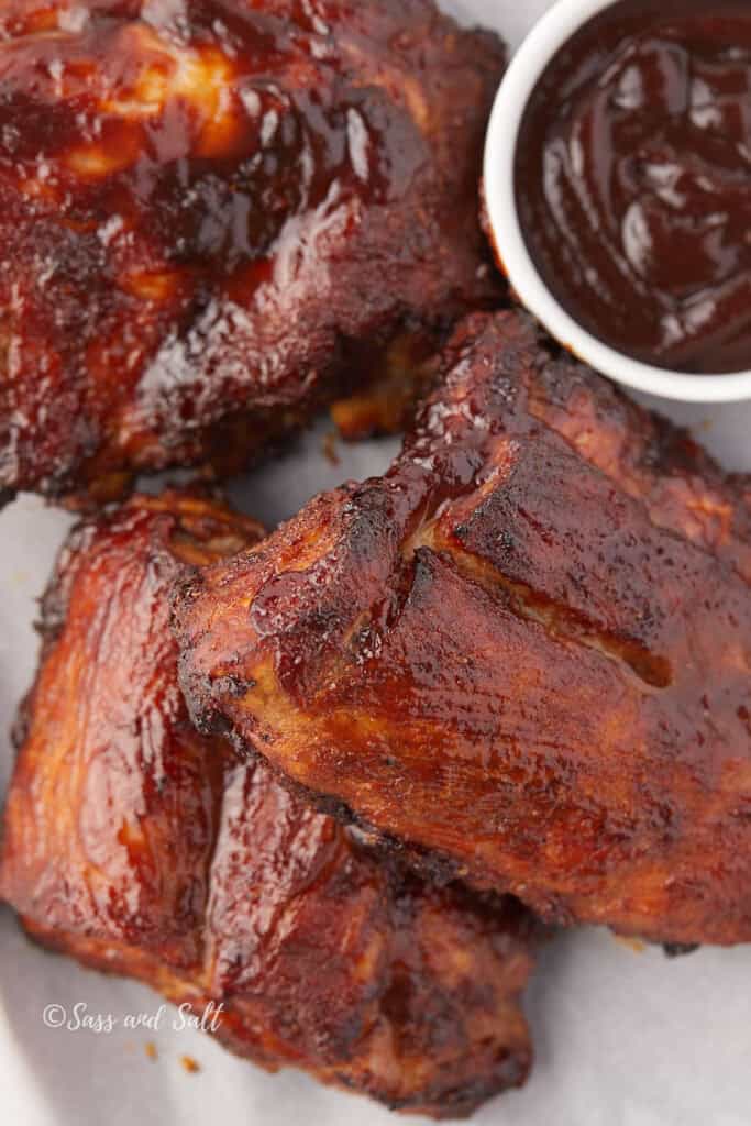 Juicy, glazed barbecued pork ribs with charred edges are stacked on a plate. A small bowl of dark, thick barbecue sauce is on the side, ready for dipping. The ribs are coated in a sticky, rich glaze.
