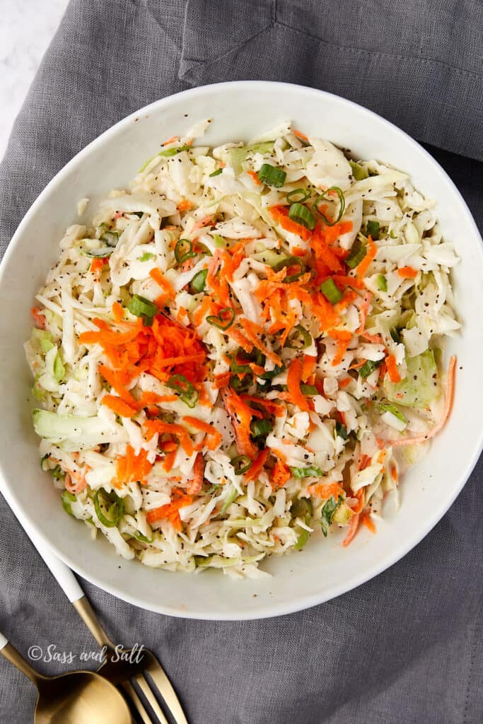 A bowl of coleslaw with shredded cabbage, green onions, and carrots on a gray napkin. Two gold forks are placed beside the bowl.