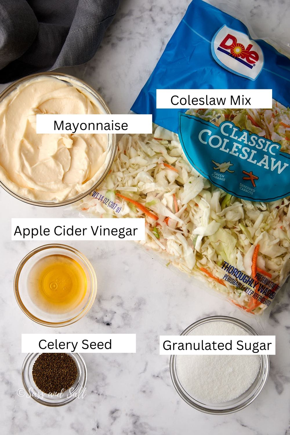 A top-view image of coleslaw ingredients on a marble surface. Items include a bag of coleslaw mix, a bowl of mayonnaise, a small bowl of apple cider vinegar, a dish with celery seed, and a bowl of granulated sugar. Labels identify each ingredient.