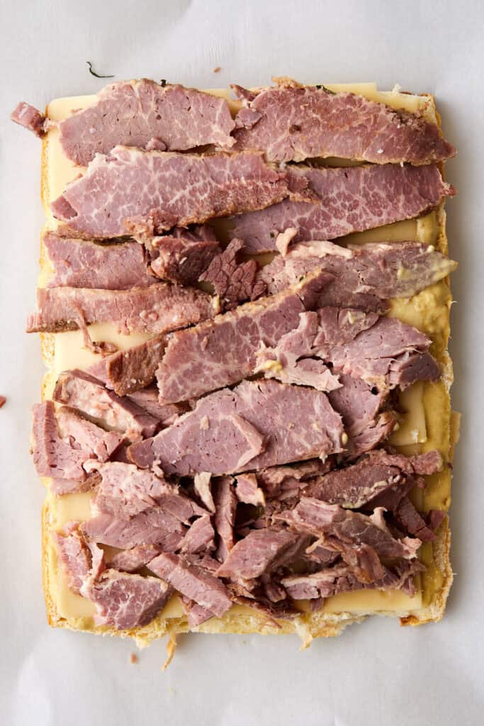 Rectangular pieces of sliced corned beef layered over melted Swiss cheese on a sheet of bread, all placed on white parchment paper.