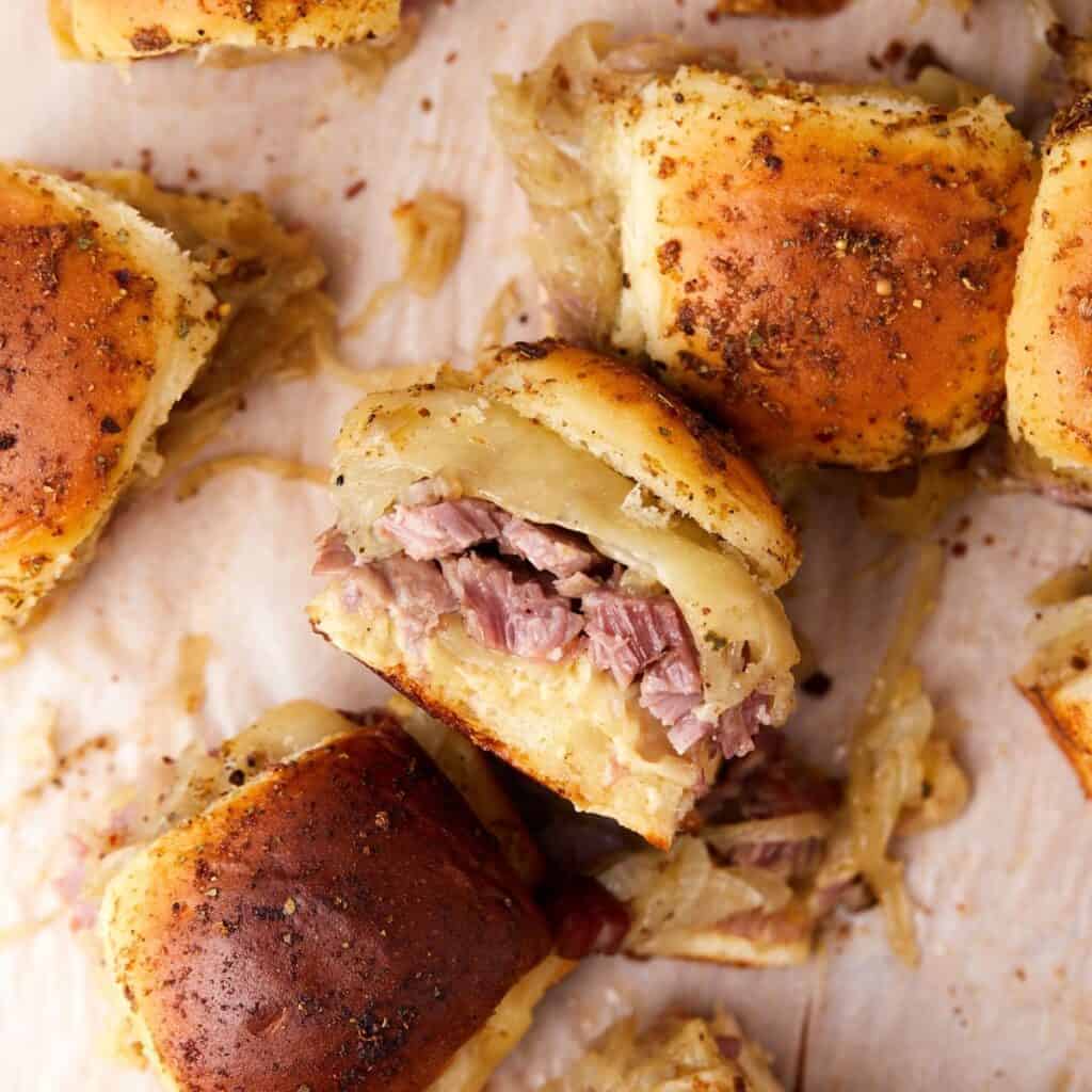 Golden toasted sliders filled with shredded meat, melted cheese, and caramelized onions are arranged on parchment paper. One slider is cut open, showing the juicy meat and gooey cheese inside.