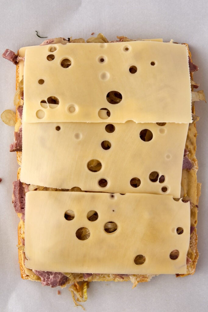 Three rectangular slices of Swiss cheese with holes are placed on top of a layer of corned beef and sautéed onions on sandwich bread. The sandwich is open-faced on a parchment-lined surface.
