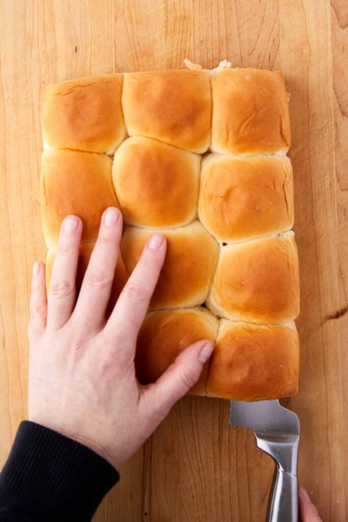 A hand rests on a tray of twelve golden brown dinner rolls on a wooden surface, while a spatula slides under one roll to lift it.