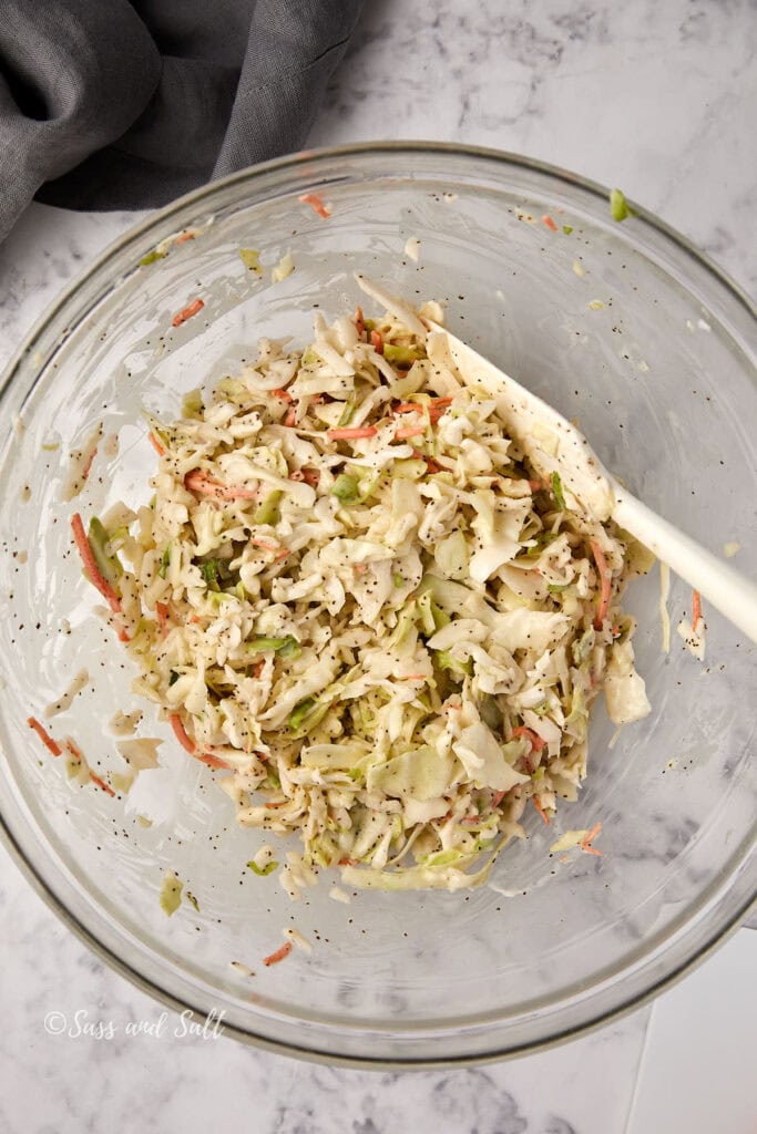 A clear glass bowl filled with coleslaw, featuring shredded cabbage, carrots, and a sprinkle of black pepper. A white spatula rests inside the bowl. The background shows a marble surface and a gray cloth in the corner.