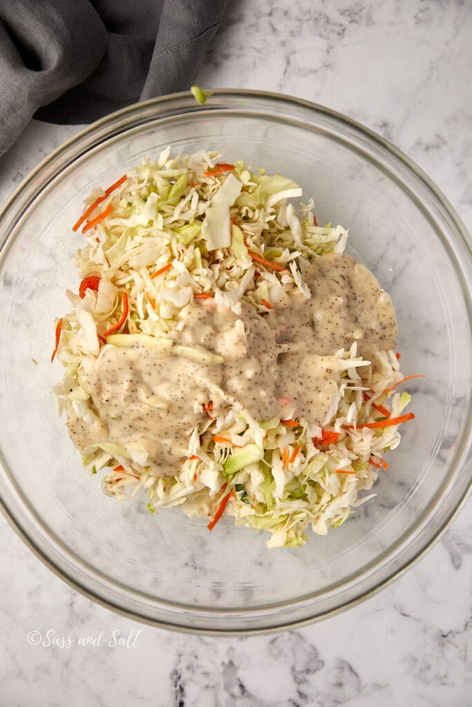 A clear glass bowl filled with chopped cabbage and carrots topped with a creamy dressing, set on a marble countertop. A gray cloth is partially visible in the top left corner.