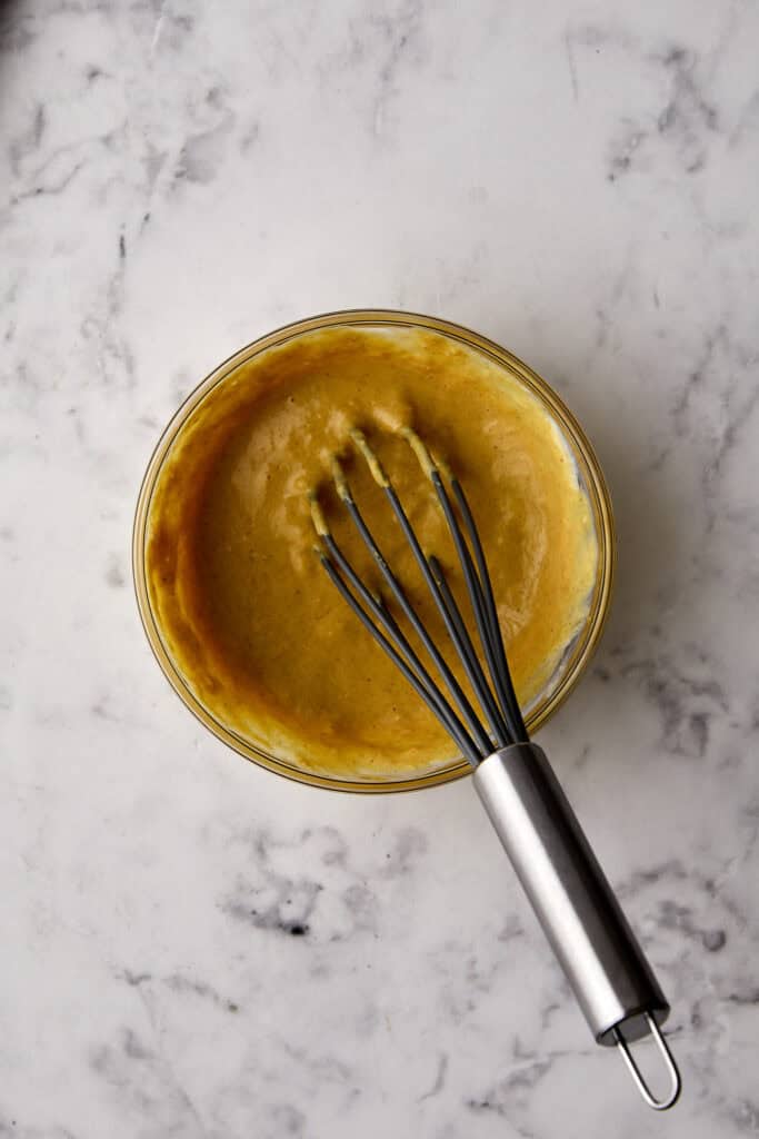 A metal whisk resting in a glass bowl filled with thick, yellow batter or sauce on a white marble surface.