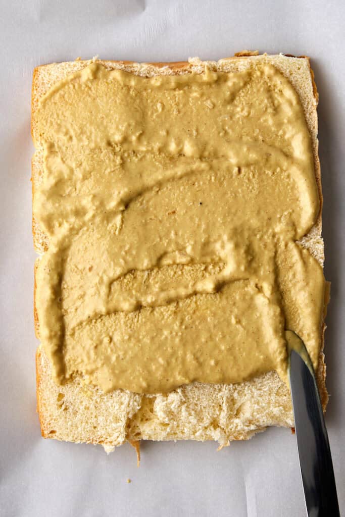 A close-up of a rectangular piece of bread being spread with a horseradish sauce using a knife. The bread rests on a sheet of parchment paper.