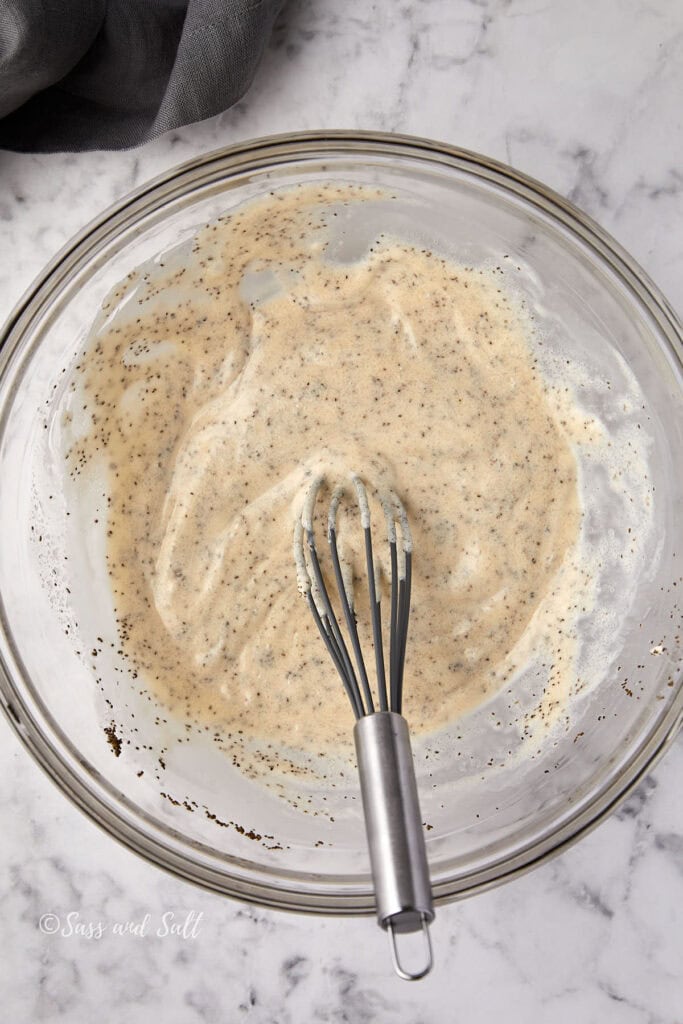 A glass bowl filled with a creamy mixture containing visible black specks, of celery seed. A metal whisk rests in the bowl. The background is a white marble surface, and a dark cloth is partially visible in the top left corner.