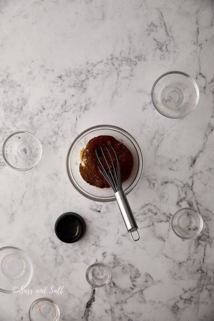 A glass mixing bowl on a marble countertop contains a variety of spices being whisked together. Surrounding the bowl are several empty glass bowls. The countertop has a white and gray marbled pattern.